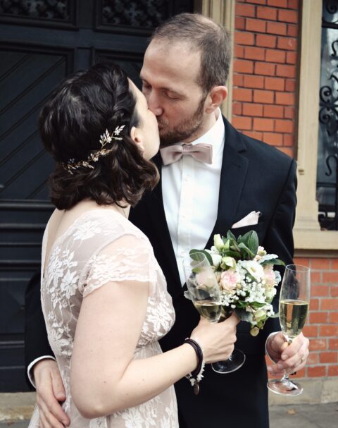Katharina Mauder und ihr frisch getrauter Ehemann küssen sich mit Sektgläsern und dem Brautstrauß in der Hand.